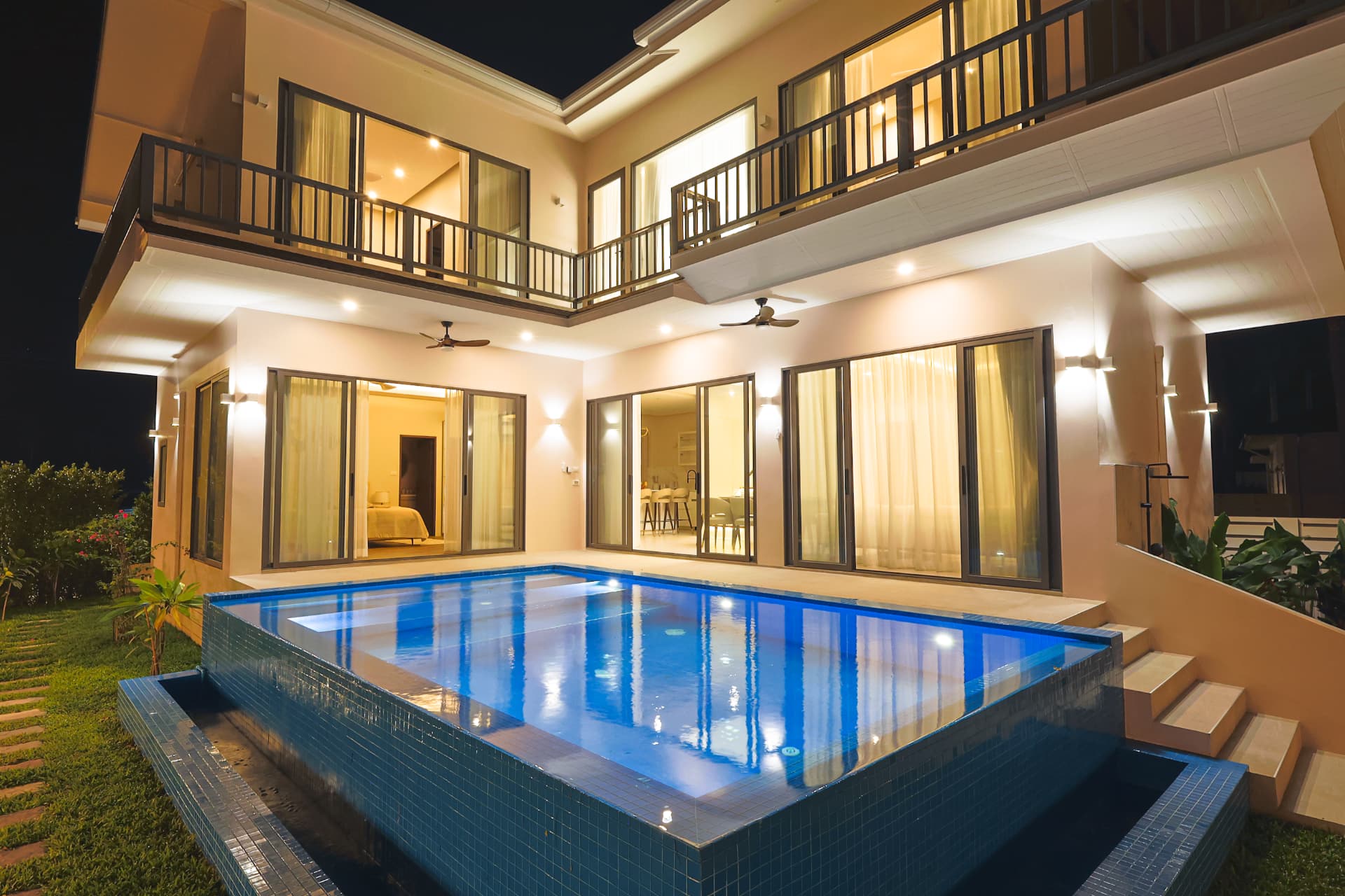 The villa lit up at night with the swimming pool in the foreground and open sliding doors on the ground floor